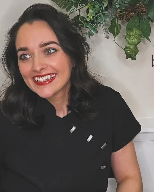 Rachel Starr, senior stylist, seated in the salon in black workwear, smiling, with greenery on the wall behind her.